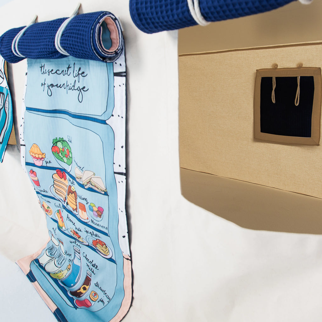 Aufrollbare Fundertable-Paneele unter dem Tisch befestigt – ein Stoffpanel mit Küchenillustration und ein Panel mit Fensteröffnung, kombiniert mit navyblauem Waffelstoff und weißen Schlaufen.