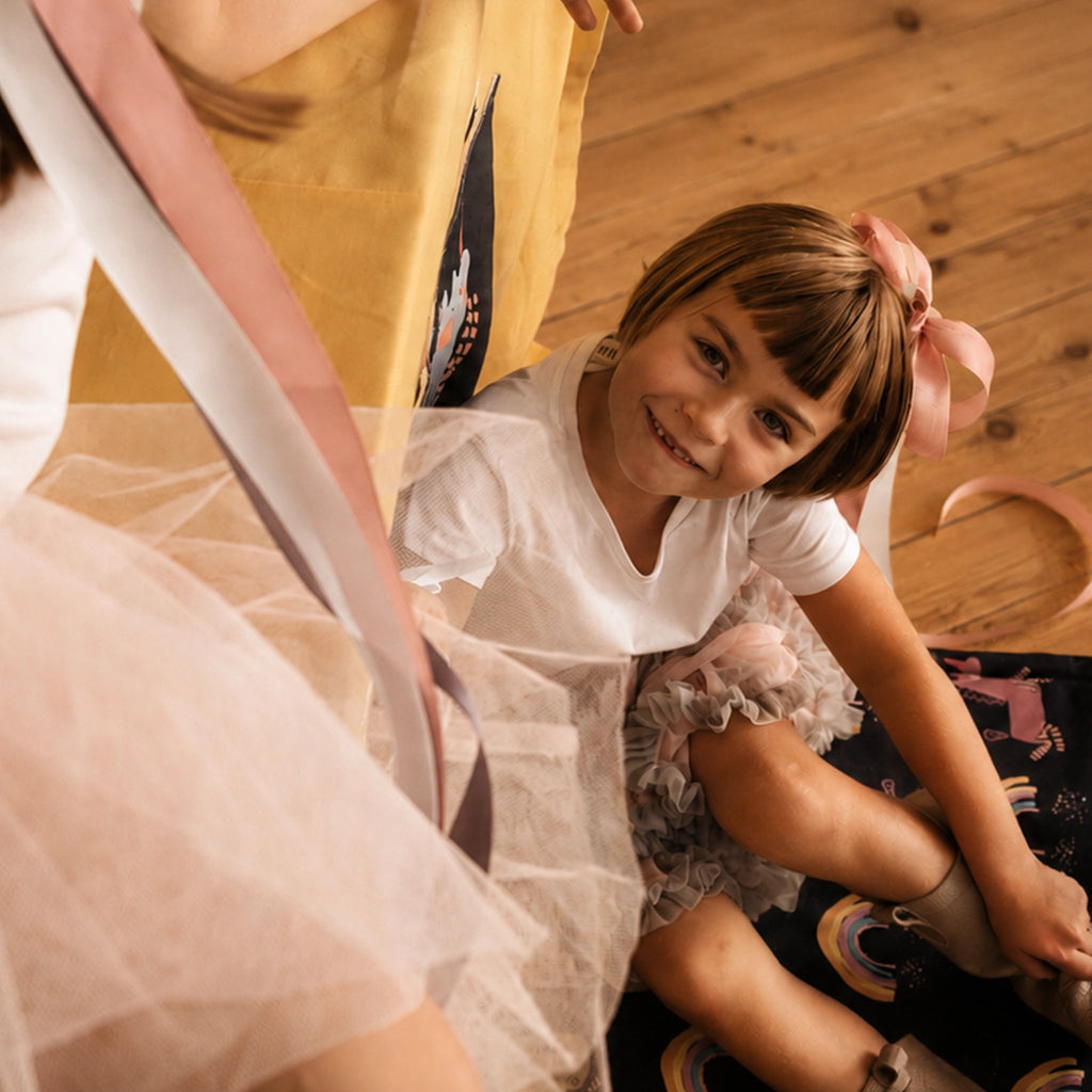 Lächelndes Kind sitzt auf einer dunklen Spielmatte mit bunten Einhörnern und Regenbögen und zieht sich Schuhe an, daneben liegt ein rosafarbenes Band