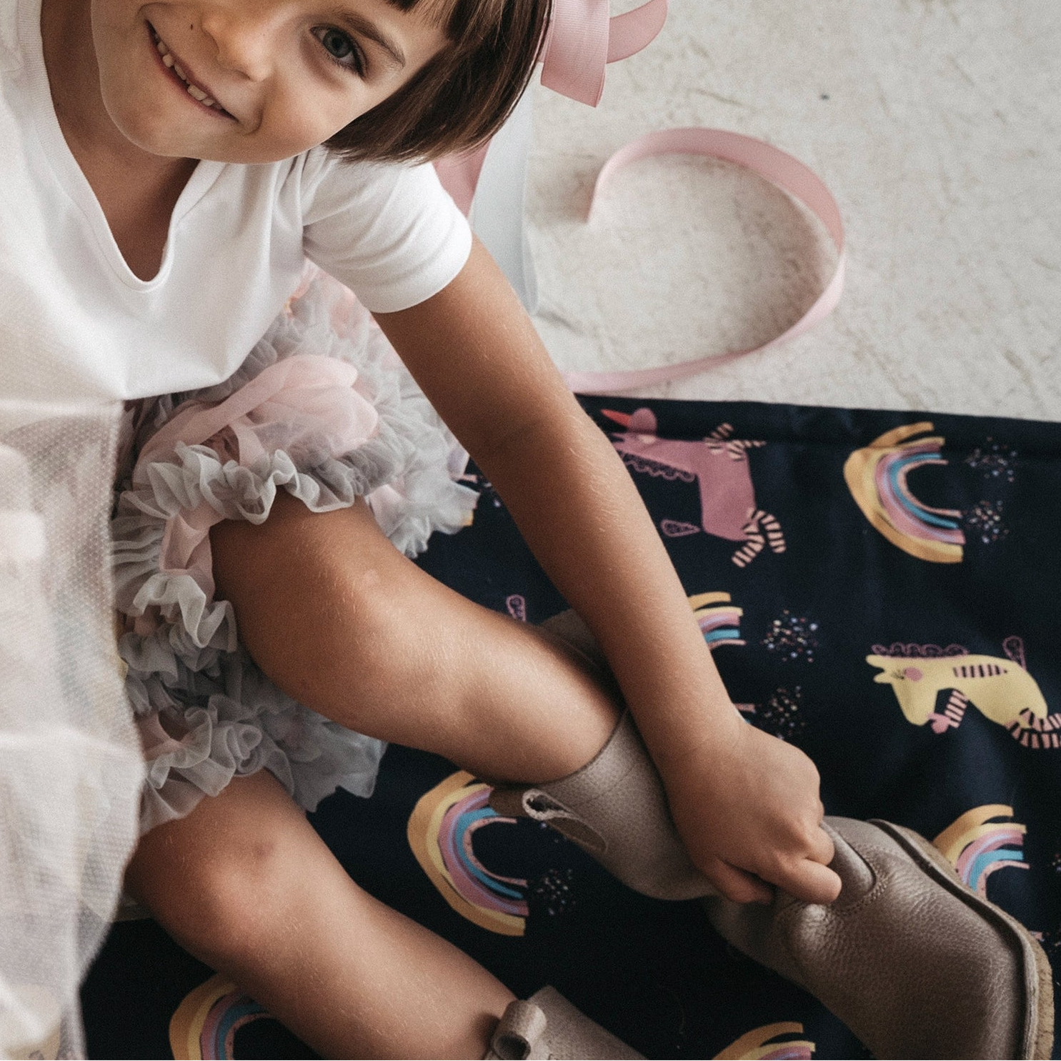 Lächelndes Kind sitzt auf einer dunklen Spielmatte mit bunten Einhörnern und Regenbögen und zieht sich Schuhe an, daneben liegt ein rosafarbenes Band