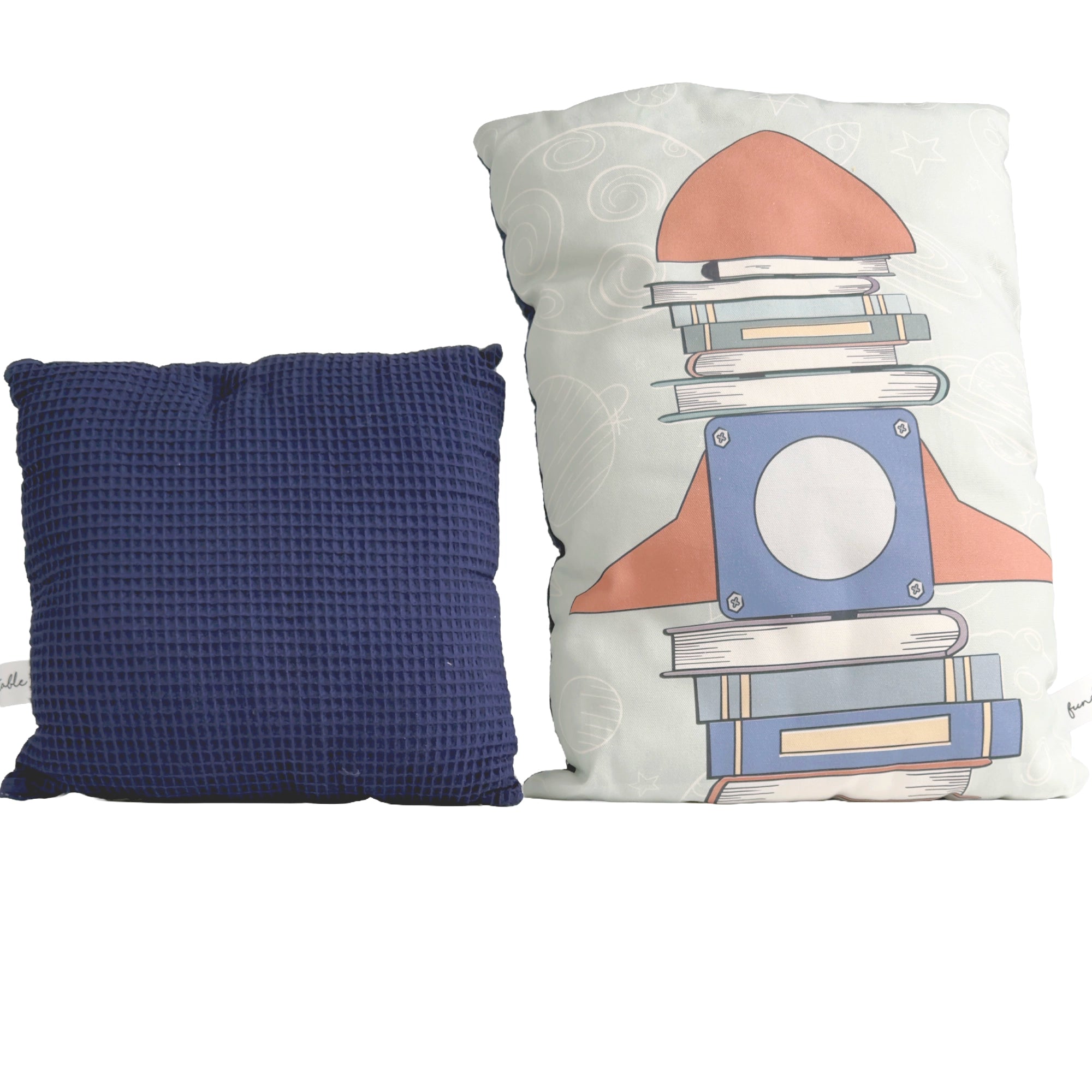 Zwei Kissen, eines mit Rakete aus gestapelten Büchern auf heller Stoffseite, das andere mit dunkelblauer, strukturierter Waffelrückseite