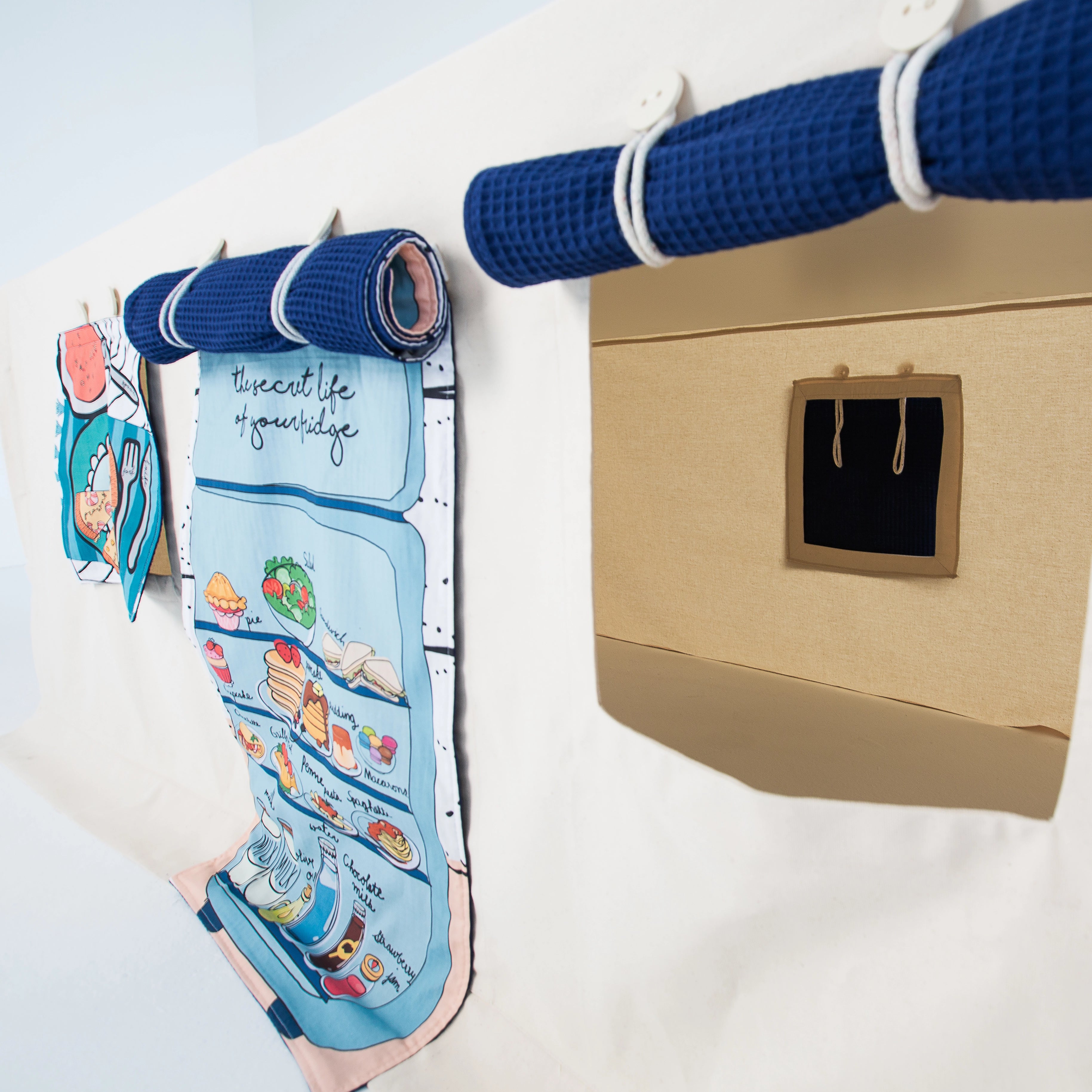 Aufrollbare Fundertable-Paneele unter dem Tisch befestigt – ein Stoffpanel mit Küchenillustration und ein Panel mit Fensteröffnung, kombiniert mit navyblauem Waffelstoff und weißen Schlaufen.