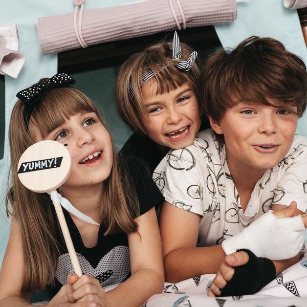 Drei Kinder lachen und spielen unter einem Fundertable Spielzelt. Ein Mädchen hält ein Holzschild mit der Aufschrift „YUMMY!“, während die Kinder gemeinsam in der gemütlichen Unter-dem-Tisch-Spielwelt sitzen.
