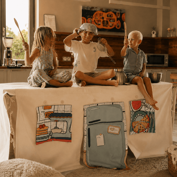 Drei Kinder sitzen auf einem Fundertable Spielzelt im Küchen-Design und spielen mit Kochutensilien. Die aufrollbaren Fensterklappen zeigen Küchenmotive wie Kühlschrank, Pizza und Backszene und schaffen eine warme, gemütliche Spielatmosphäre im Familienalltag.