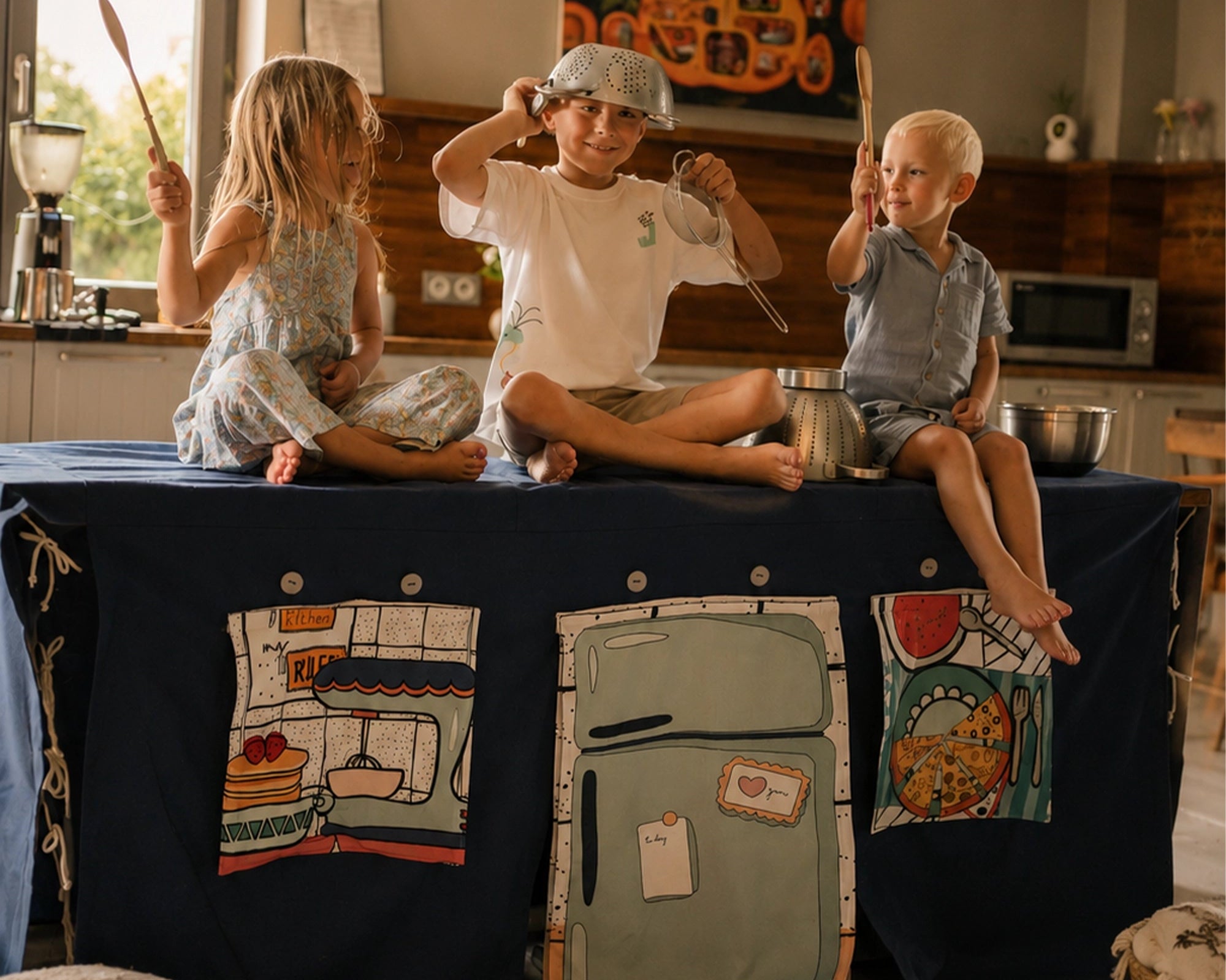 Drei Kinder sitzen auf einem Fundertable-Spielhaus in einer Küche und spielen mit Kochutensilien. Dunkelblaue Base mit Küchenklappen und Kühlschrank-Tür im warmen Abendlicht.
