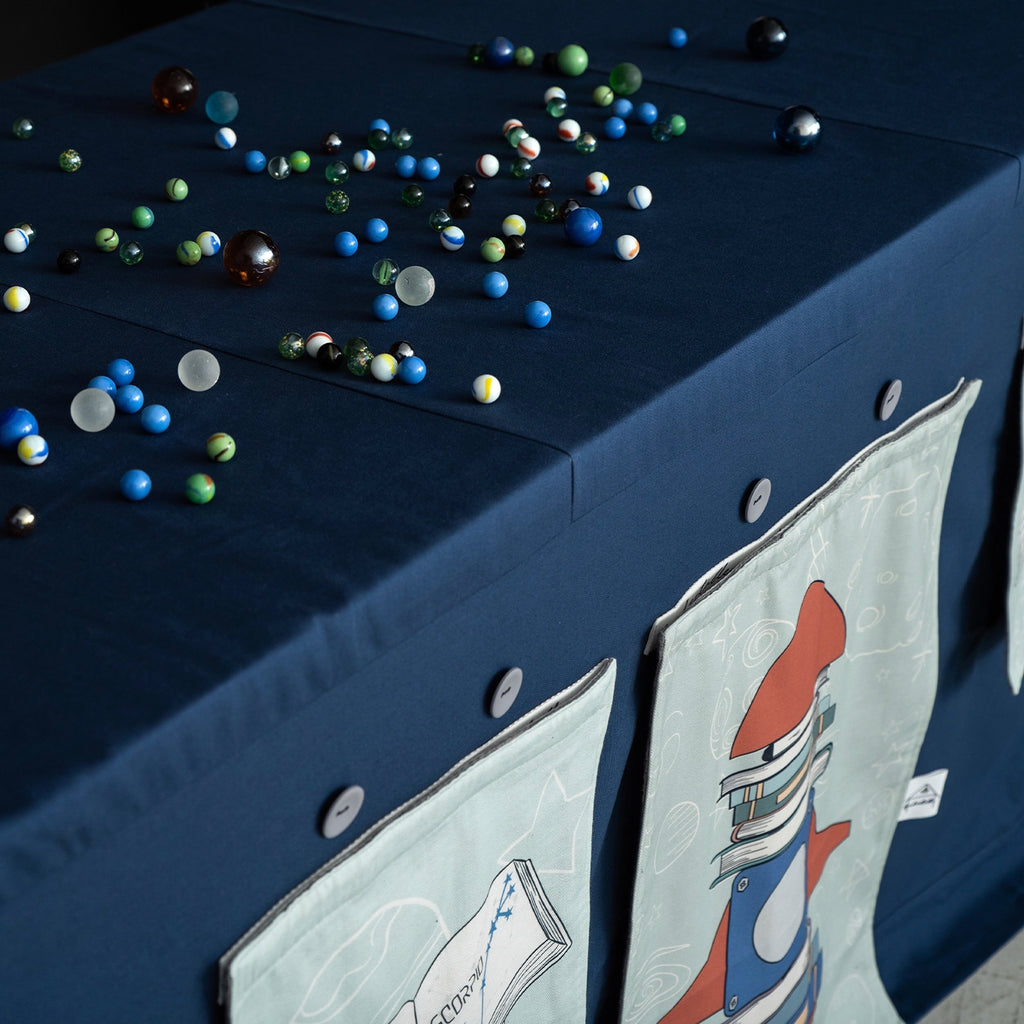 Table covered with a fabric play tent featuring illustrated window panels and small toys on top