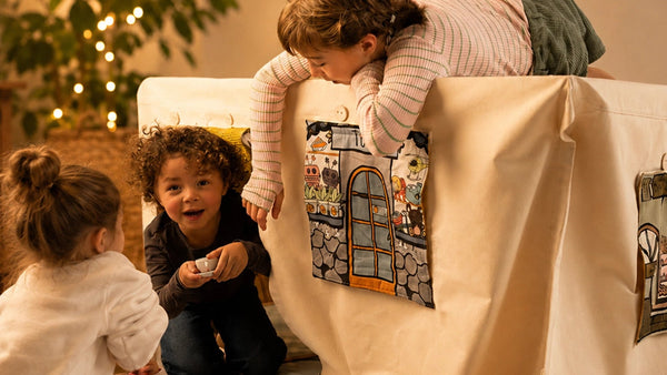 Kinder spielen rund um ein helles Fundertable Spielzelt mit aufrollbaren Fensterklappen im Funderville-Design. Ein Kind schaut von oben in die Spielwelt hinein, während andere darunter mit Spielgeschirr und Rollenspiel spielen.
